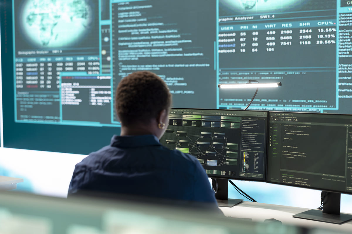 A auditoria de logs é realizada por uma profissional de TI vista de costas, focada em múltiplos monitores que exibem dados, códigos e gráficos complexos de sistema.