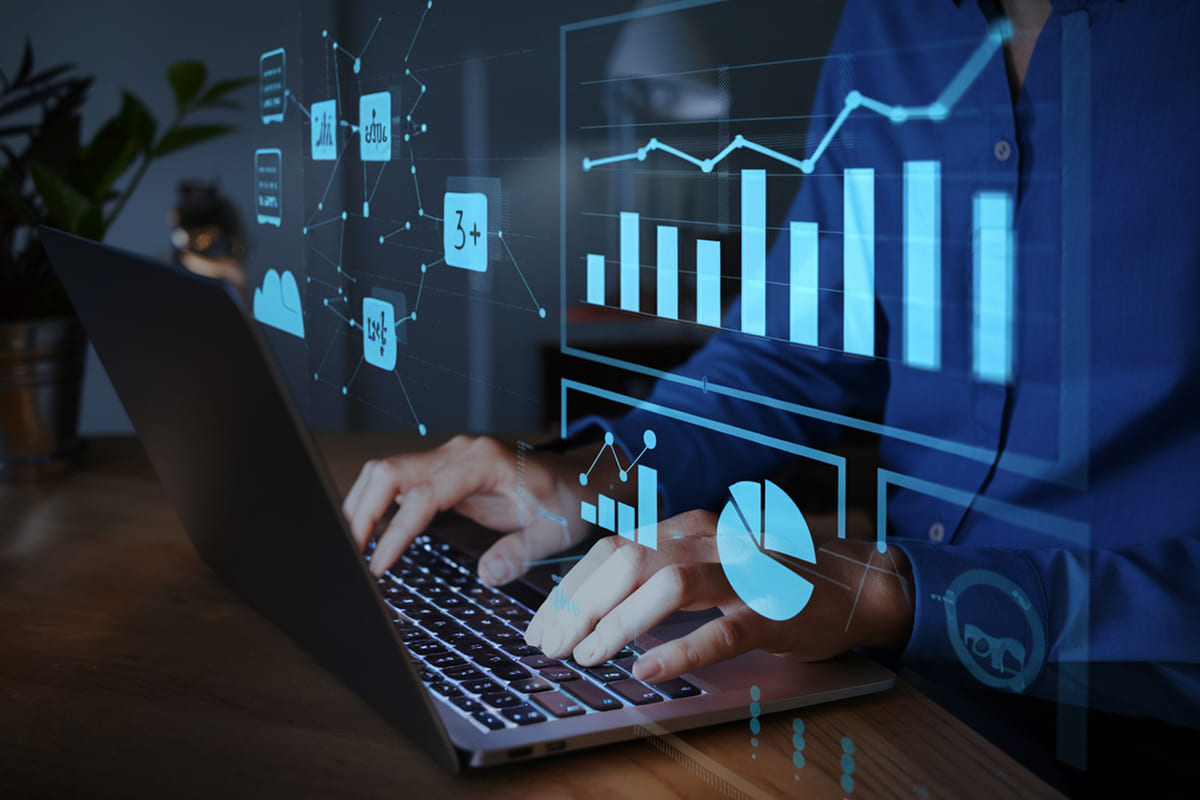 A prática da data literacy é representada por mãos digitando em um notebook no escuro, com projeções luminosas de gráficos, estatísticas e painéis de dados azuis flutuando à sua frente.