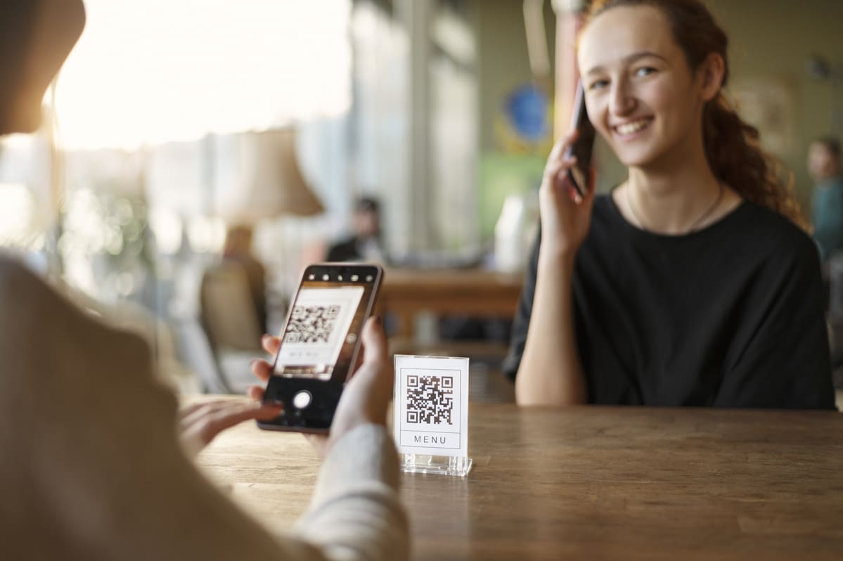 A experiência figital na prática com uma cliente escaneando o cardápio em QR code pelo celular em uma mesa de café ao lado de uma amiga sorridente.